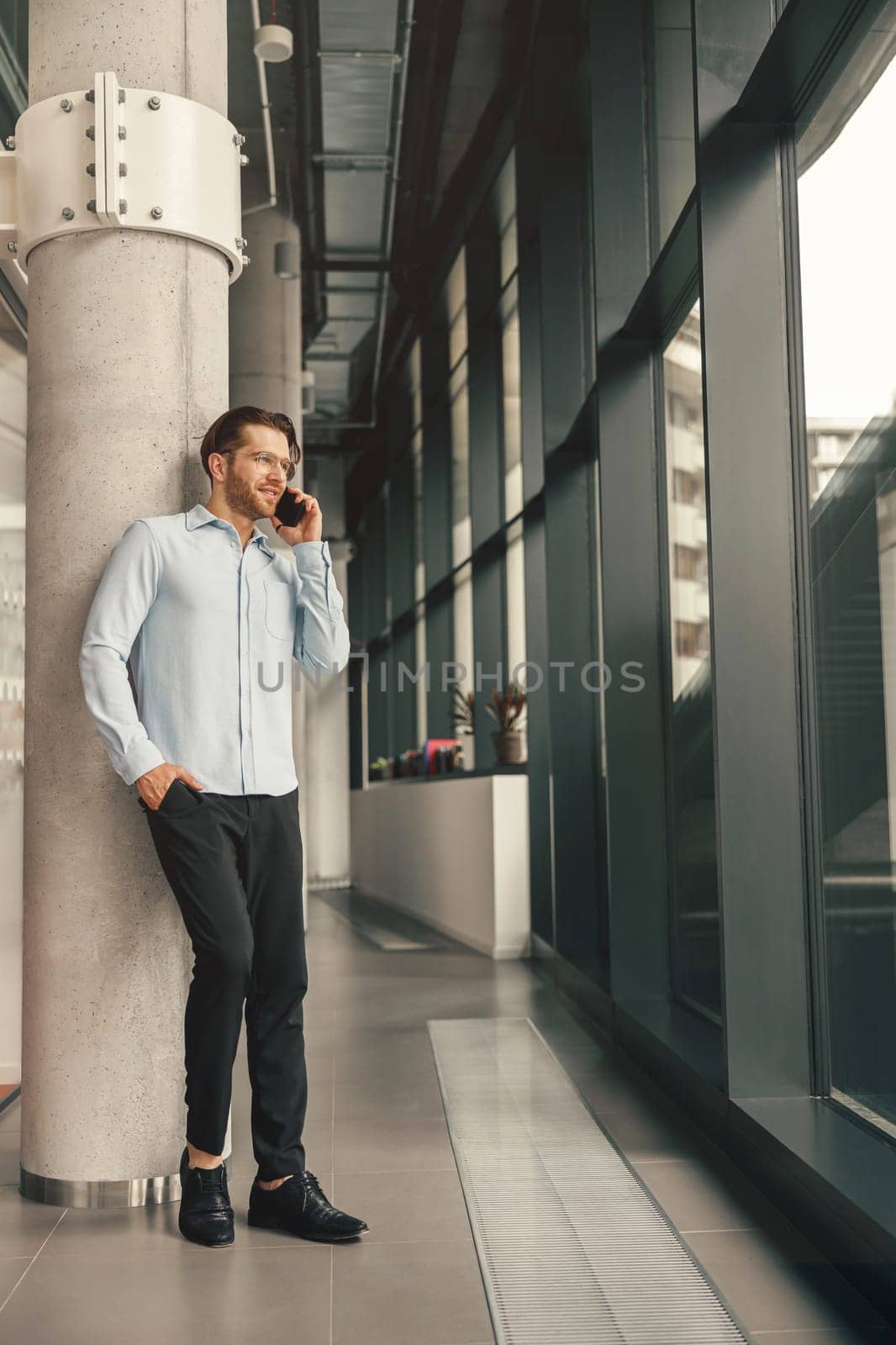 Focused male entrepreneur talking phone with client while standing in office and looking at side by Yaroslav_astakhov