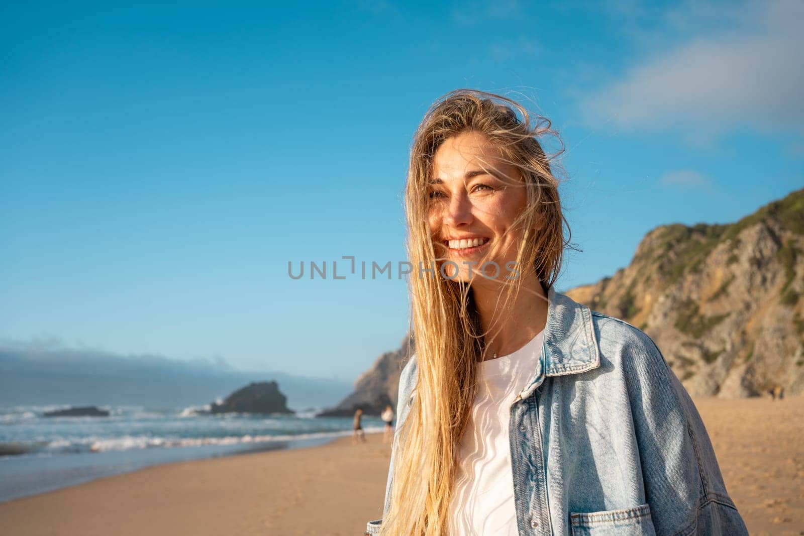 Woman with happy smile on beach. Portrait of female smiling at sea shore with huge rocks on background. Blond woman stand on sandy beach and has radiant smile on her face. Vacation Traveling concept