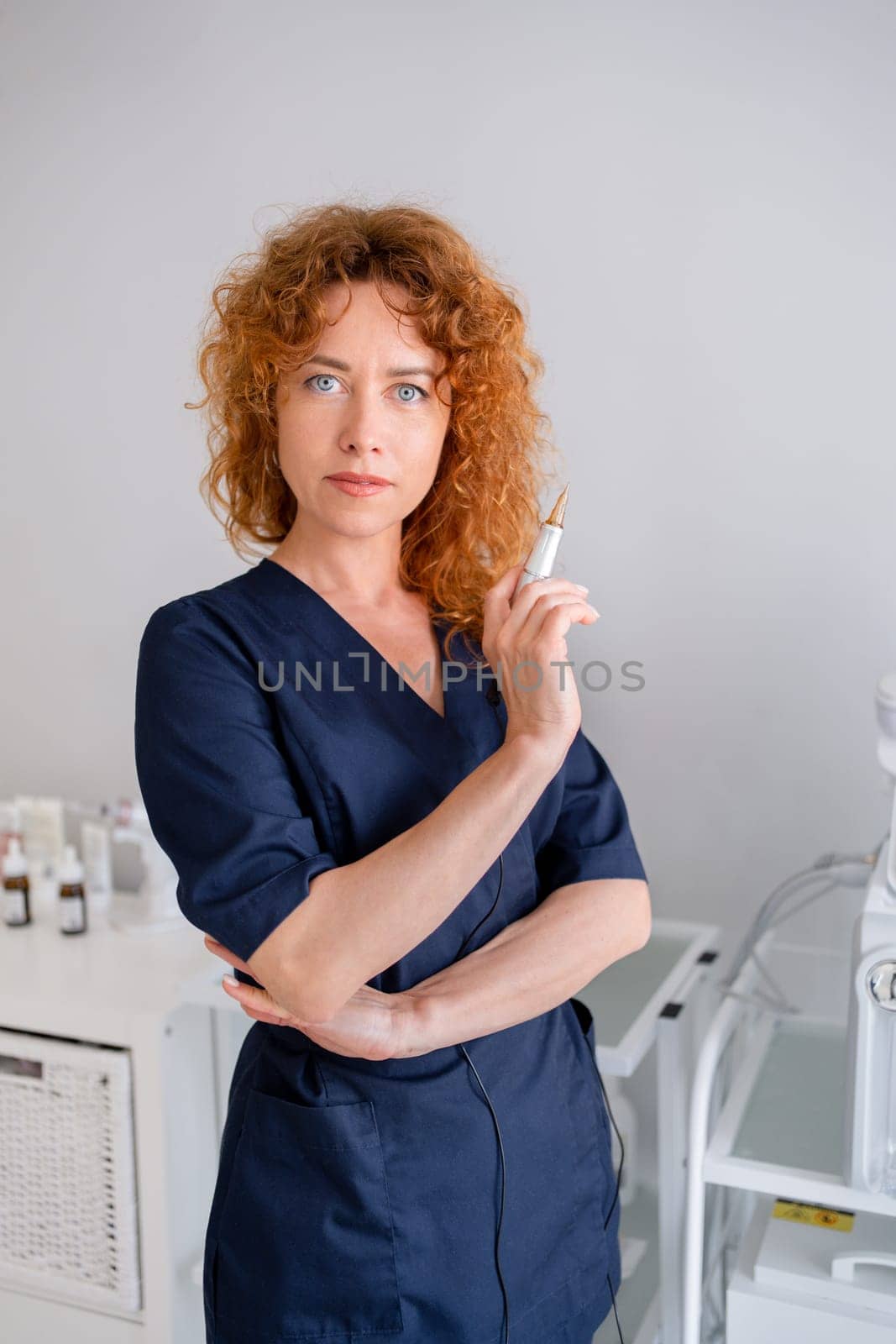 Female cosmetologist at medical office. Redhead woman dermatologist wearing scrubs holds an ampule at hospital. Skin care practitioner in navy blue medical uniform stands in aesthetic clinic office