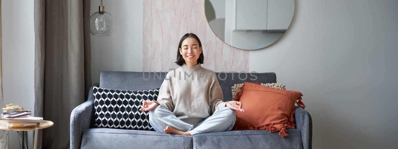 Relaxation and patience. Smiling young asian woman in cozy room, sitting on sofa and meditating, doing yoga mindfulness training by Benzoix