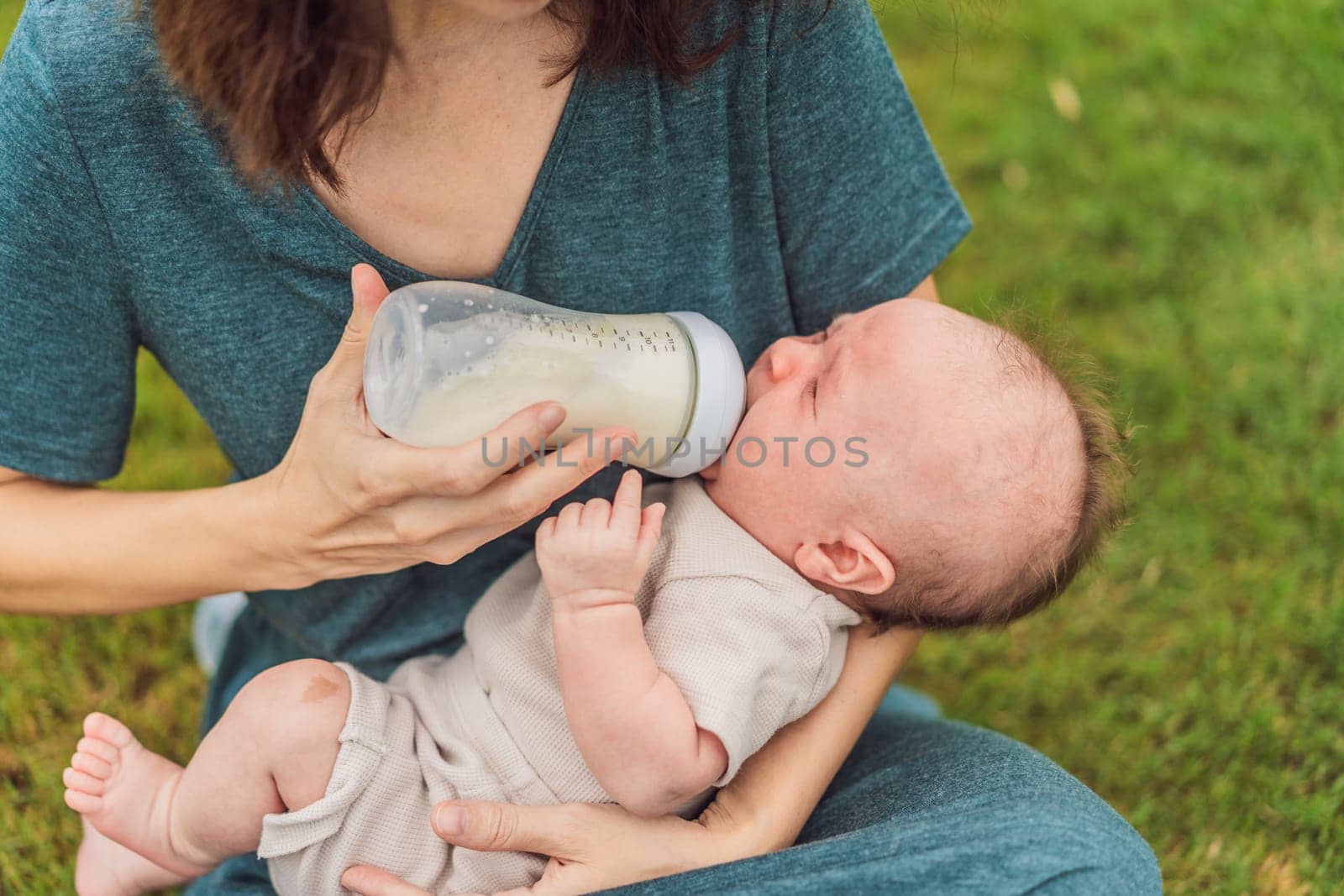 Mother holding and feeding baby from milk bottle in the park. Portrait of cute newborn baby being fed by her mother using bottle. Loving woman giving to drink milk to her boy by galitskaya