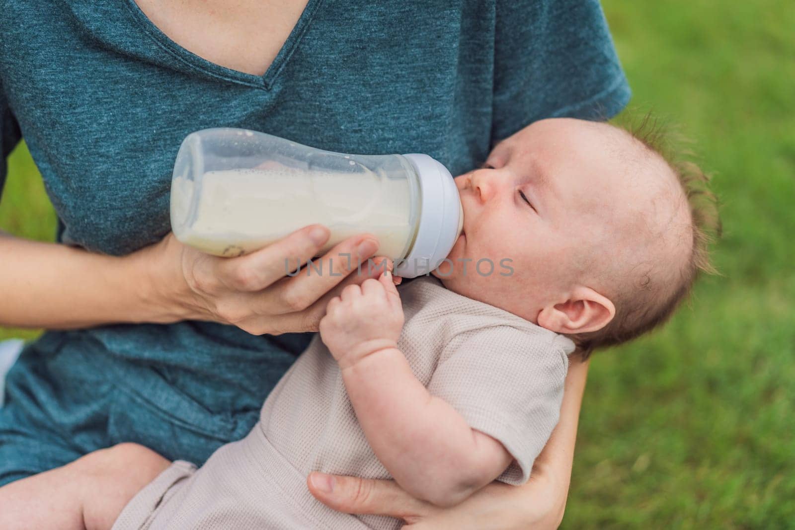 Mother holding and feeding baby from milk bottle in the park. Portrait of cute newborn baby being fed by her mother using bottle. Loving woman giving to drink milk to her boy.