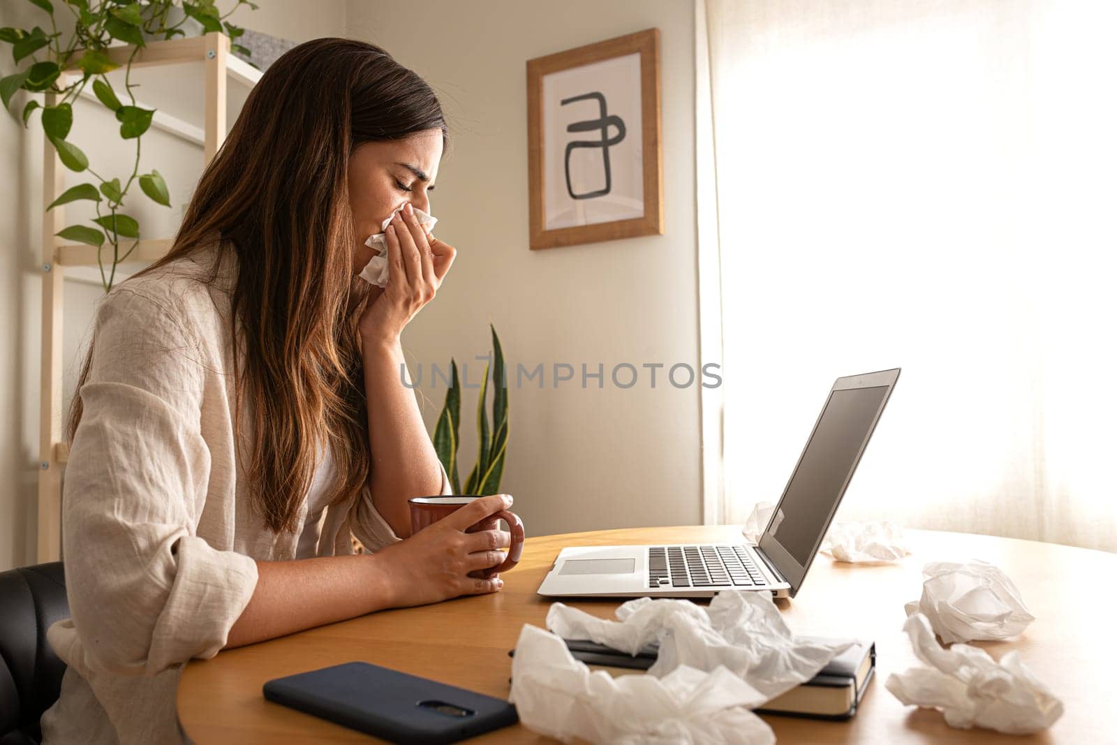 Side view of sick woman working from home office. Caucasian female blowing nose with tissue while working with laptop. Copy space. Wellness concept.