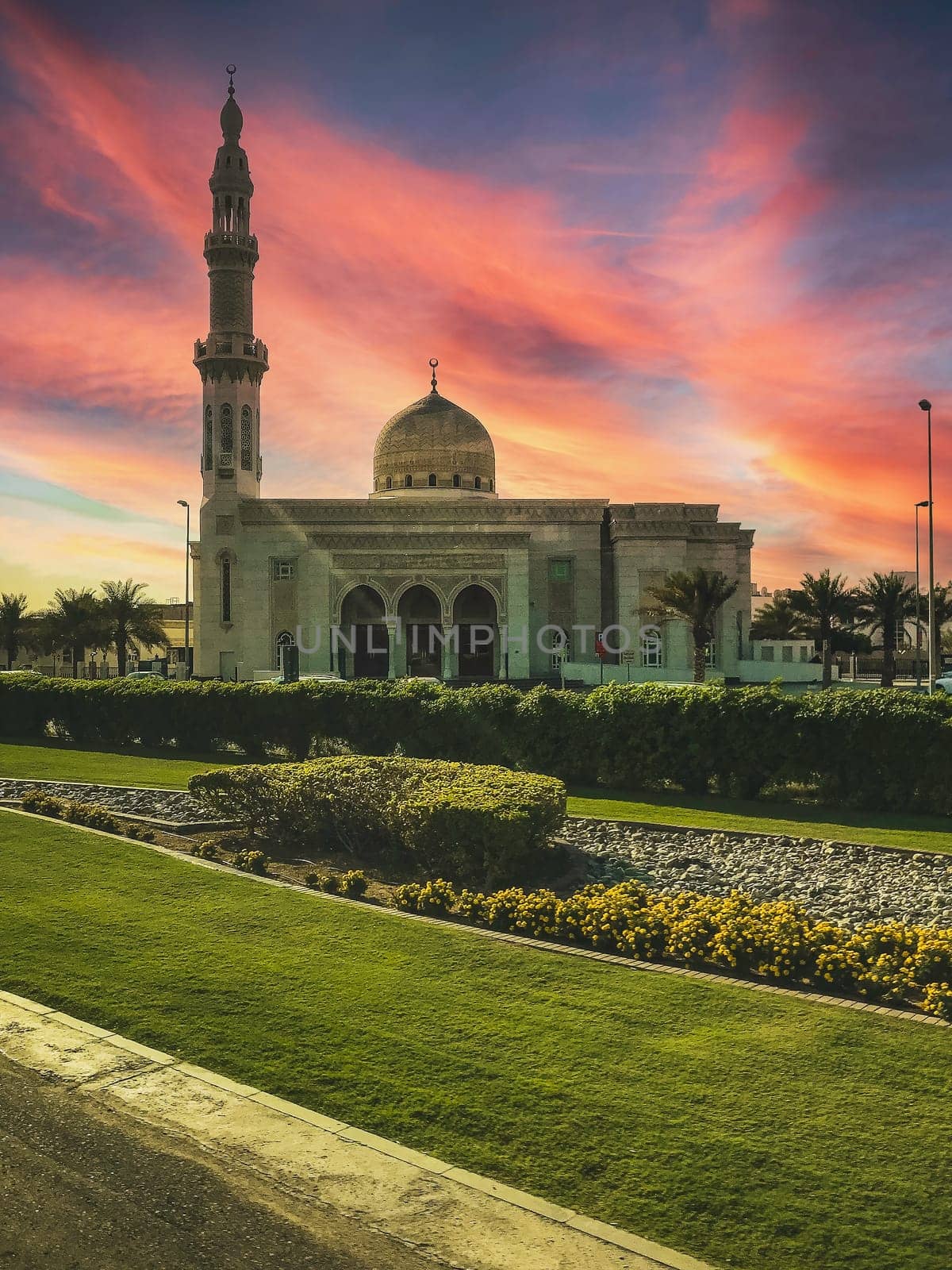 A photo of a white mosque with a dome and a minaret at sunset. The mosque is surrounded by a green garden with yellow flowers. The image has a low angle and an orange and pink sky. High quality photo