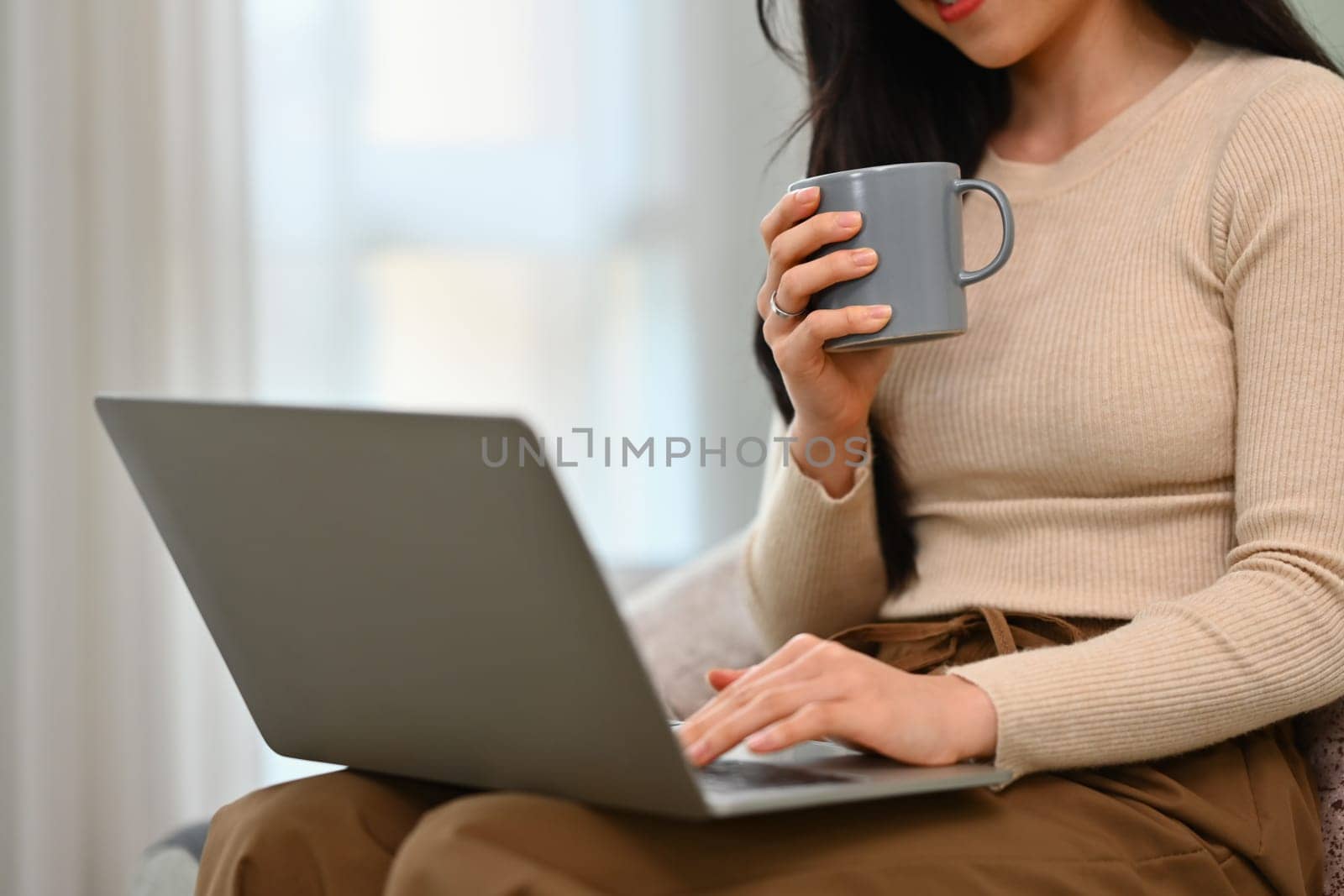 Young woman in casual clothes drinking hot tea and using laptop on couch. People, technology and domestic life.