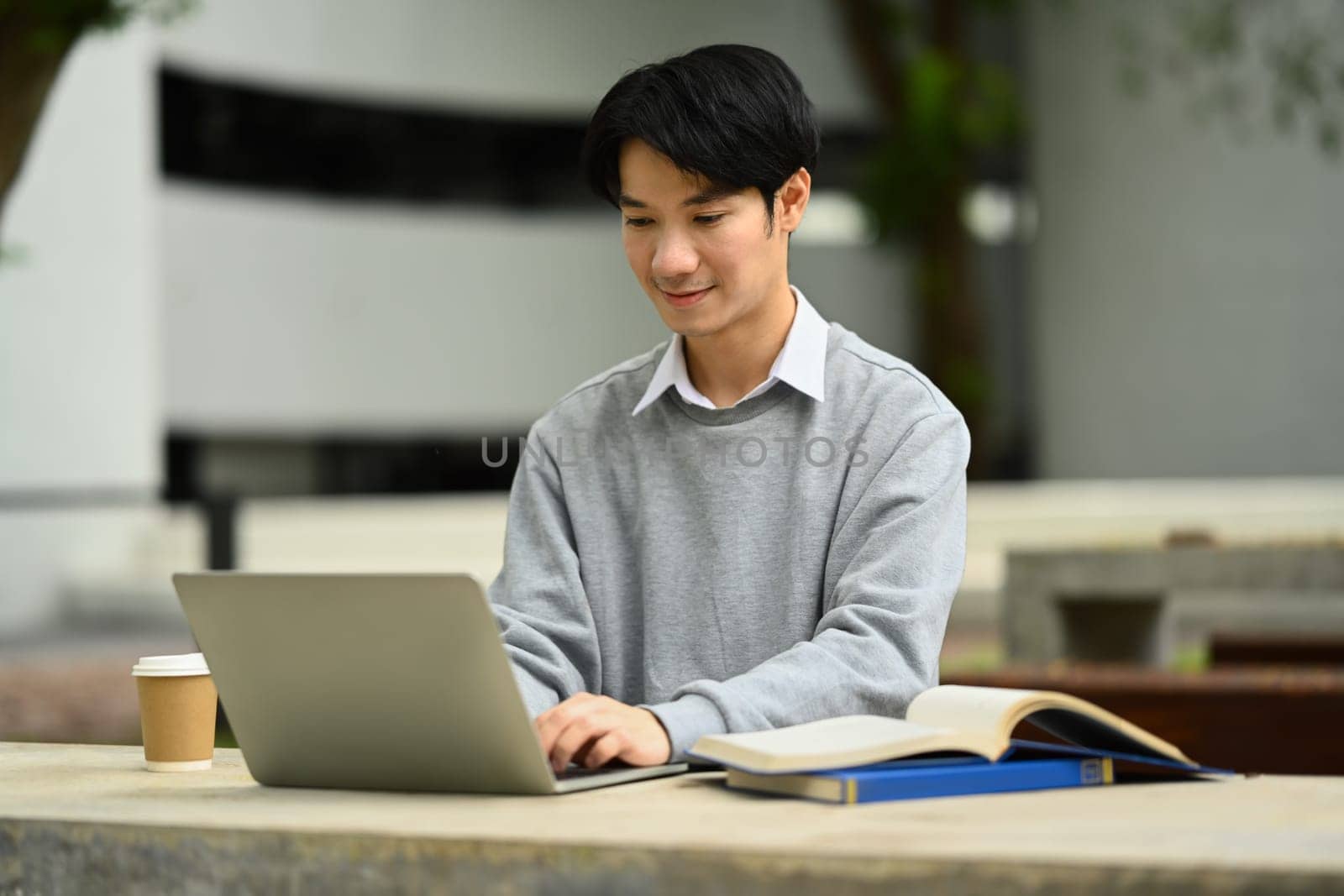 Happy asian student man typing on laptop, searching information, surfing the internet while sitting at park table.