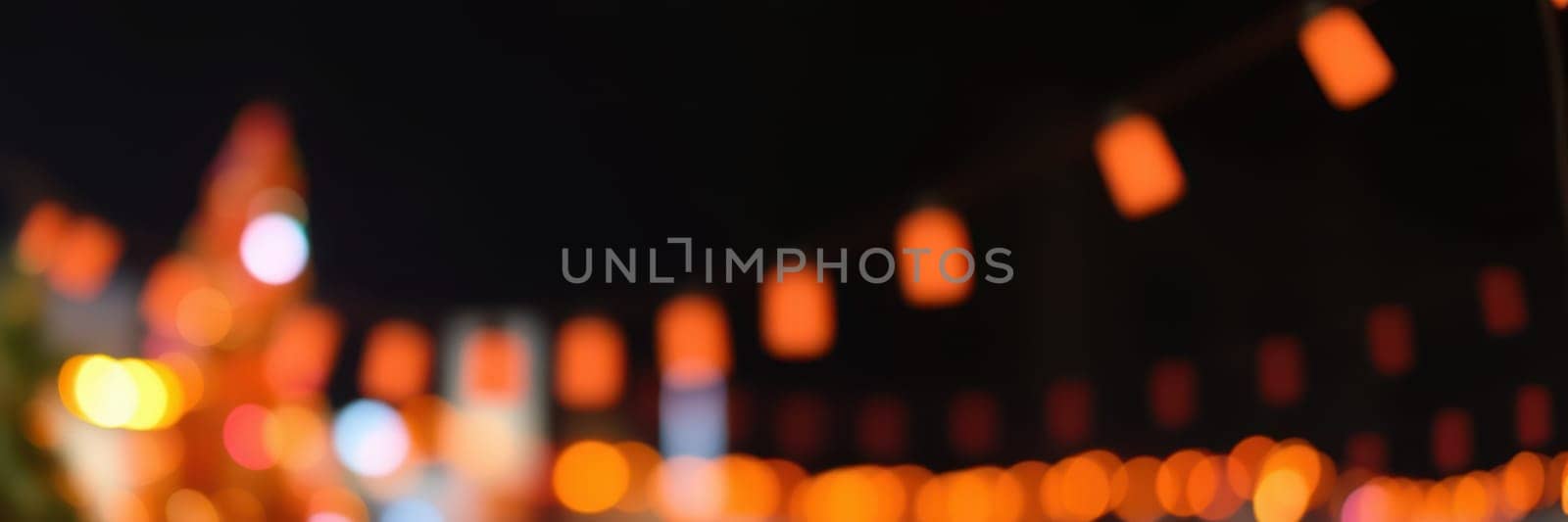 Vibrant orange lights hang over a lively night market, creating a festive atmosphere. The background features blurred city buildings and bustling activity, enhancing the celebrations.