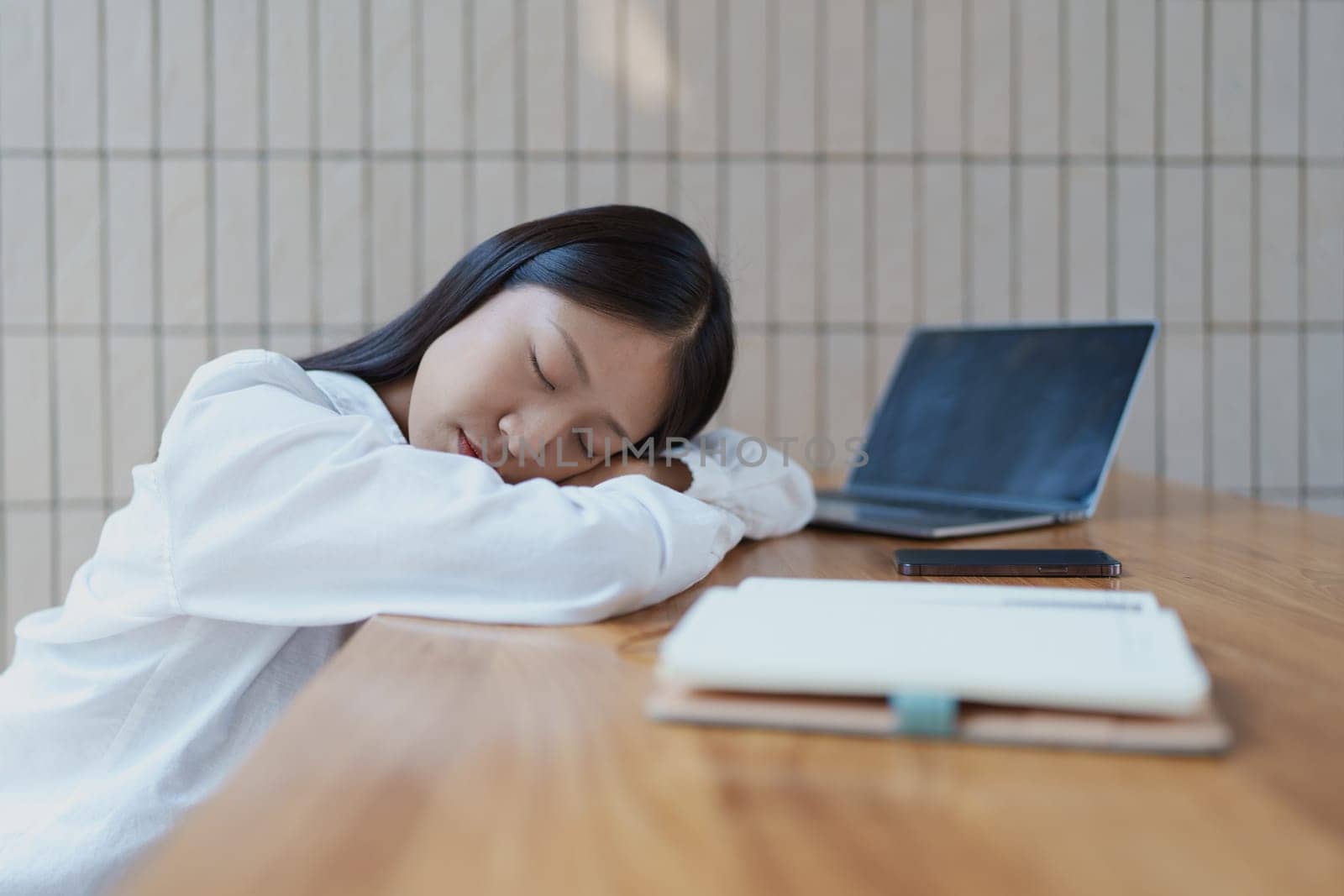 A young woman rests her head on her arms at a desk, feeling overwhelmed. The laptop beside her reflects a modern workspace, highlighting the need for rest and balance in daily life.