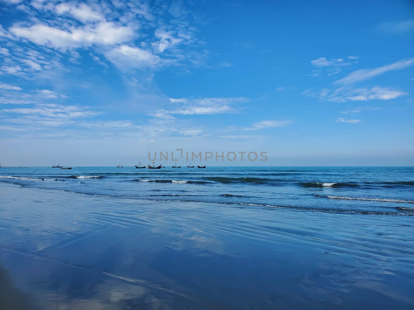 A calm beach scene with clear skies and gentle waves. Small fishing boats float in the distance on a blue ocean.