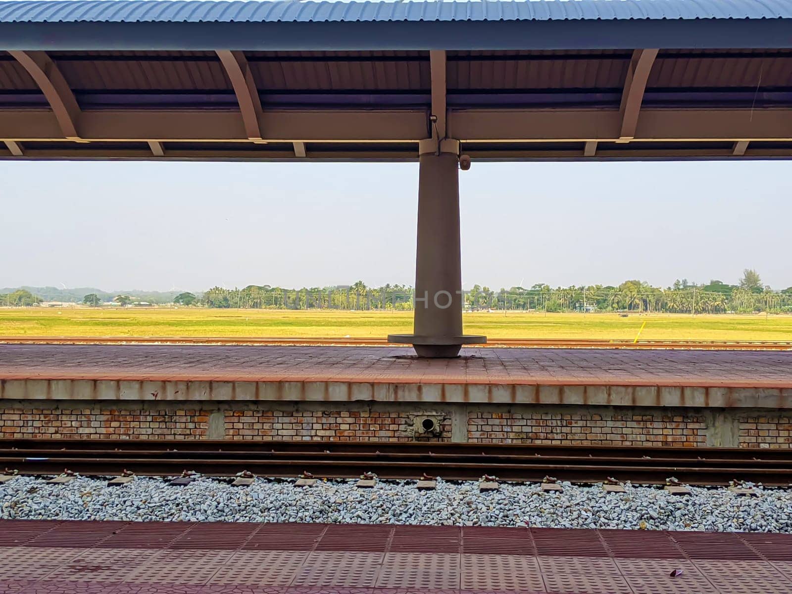 The image shows an empty train platform with a covered roof, surrounded by an open field with sparse trees in the distance.
