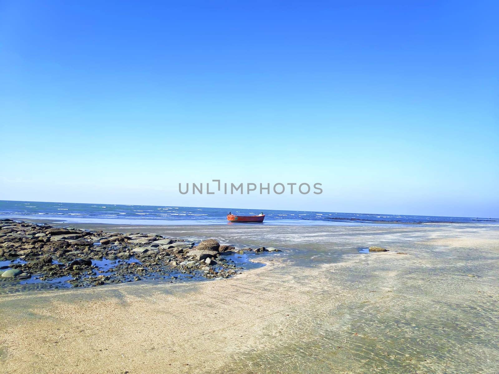 A red boat is stranded on a shallow, rocky shoreline with clear water and a vast, calm sea under a blue sky.