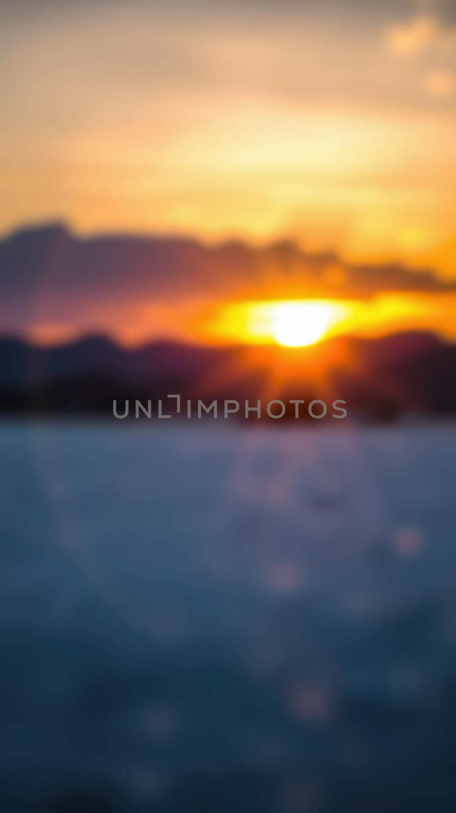 A picturesque sunset casts warm golden and orange shades across the sky, silhouetting distant mountains. The tranquil sea reflects the vibrant colors, blending with a soft blur.