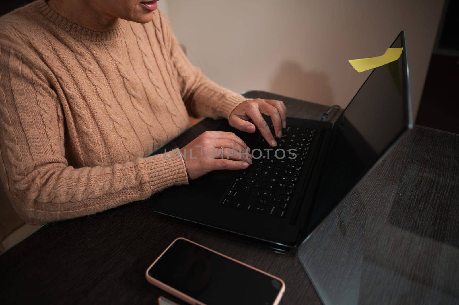 A close-up of a person wearing a warm sweater, working on a laptop at a wooden table. The setting suggests a home office or study area, with a smartphone nearby.