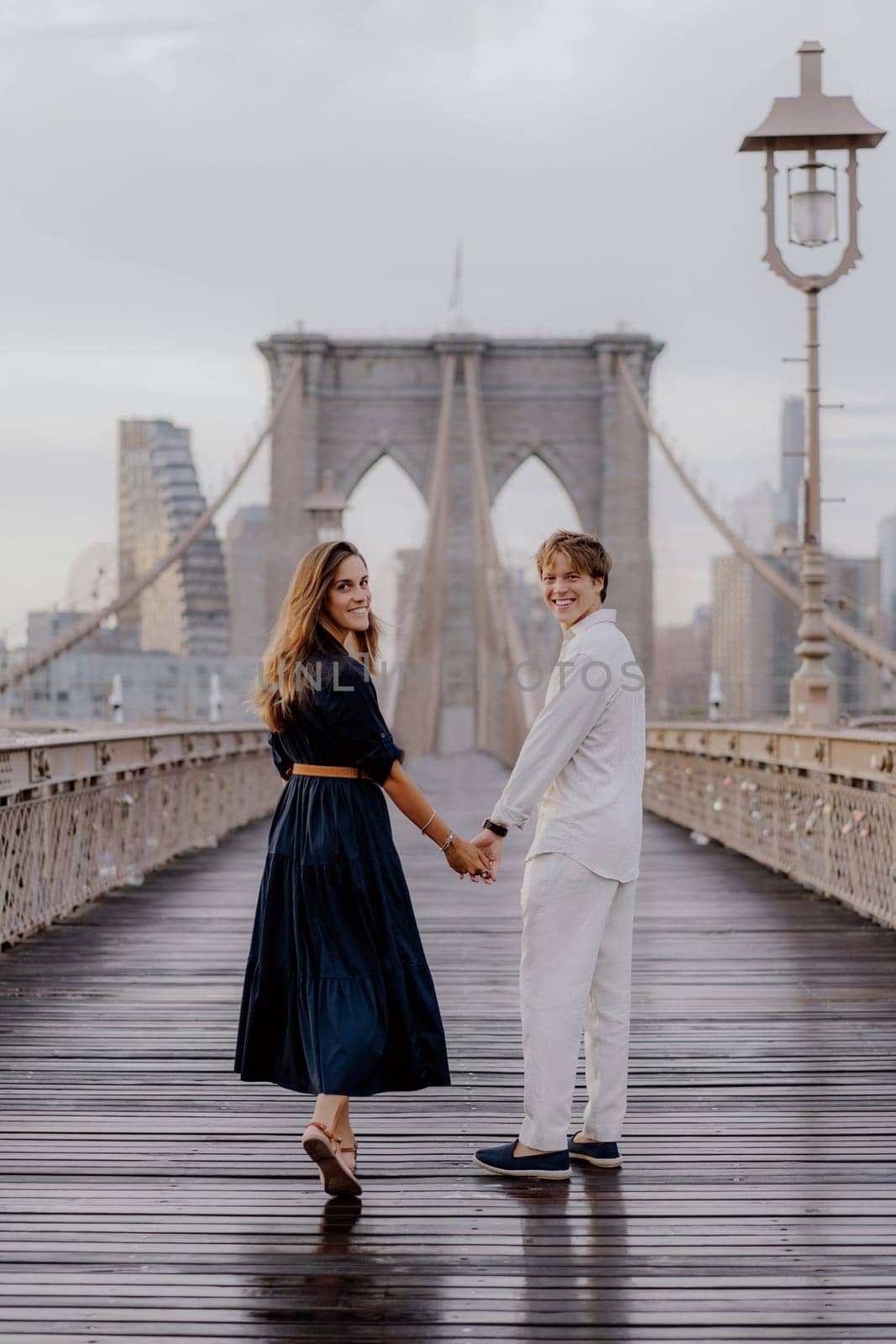 A couple strolls hand in hand on Brooklyn Bridge, surrounded by a stunning skyline and beautiful morning light