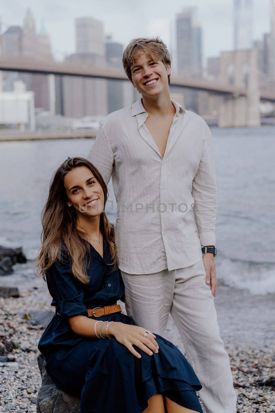 A fashionable couple joyfully shares a moment by the picturesque water, with a breathtaking city skyline behind them. Brooklyn bride and Manhattan in New York.