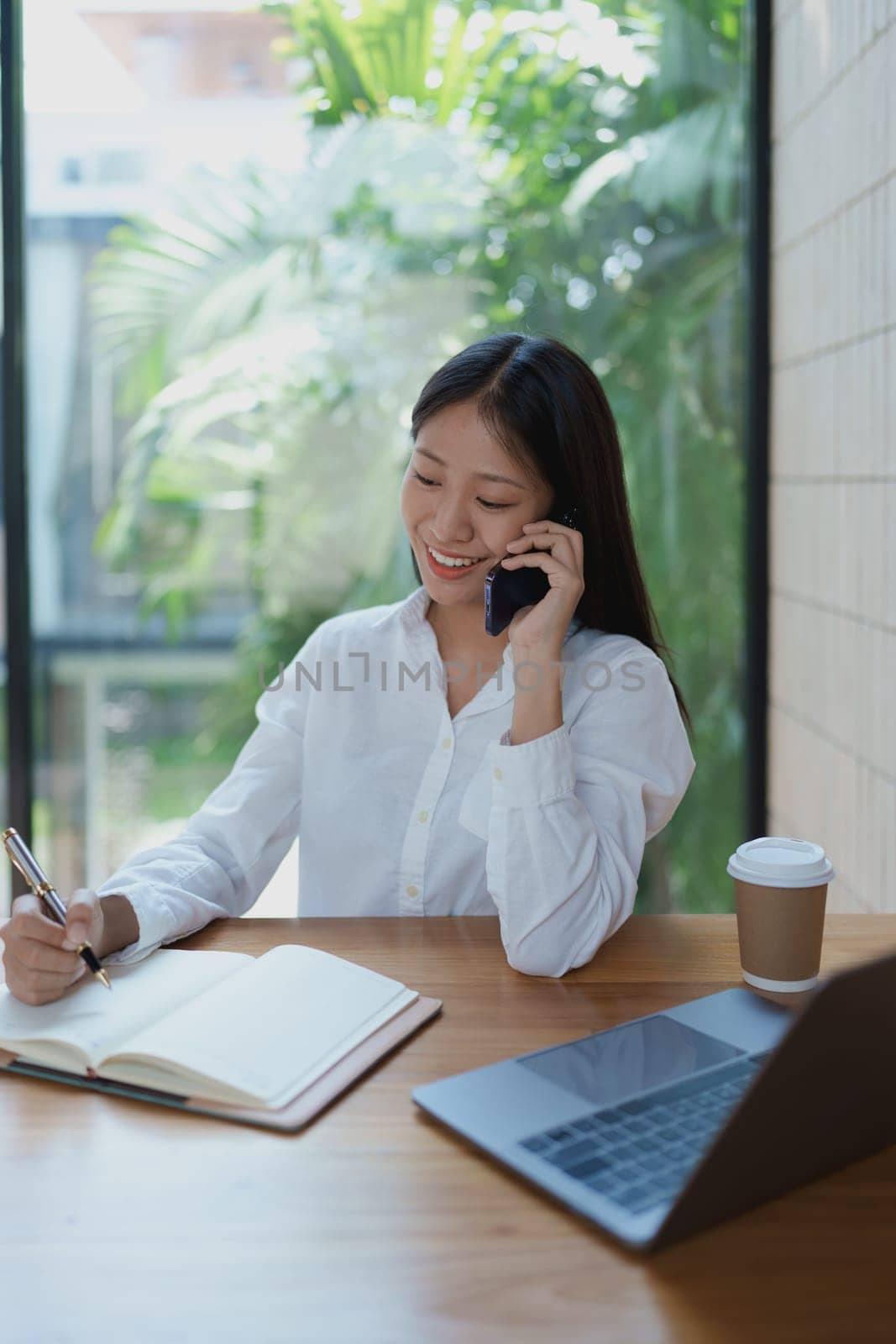 A young woman smiles while talking on the phone, jotting down notes in her notebook. A sleek laptop and coffee cup rest on the wooden table, creating an inviting workspace with greenery in view.