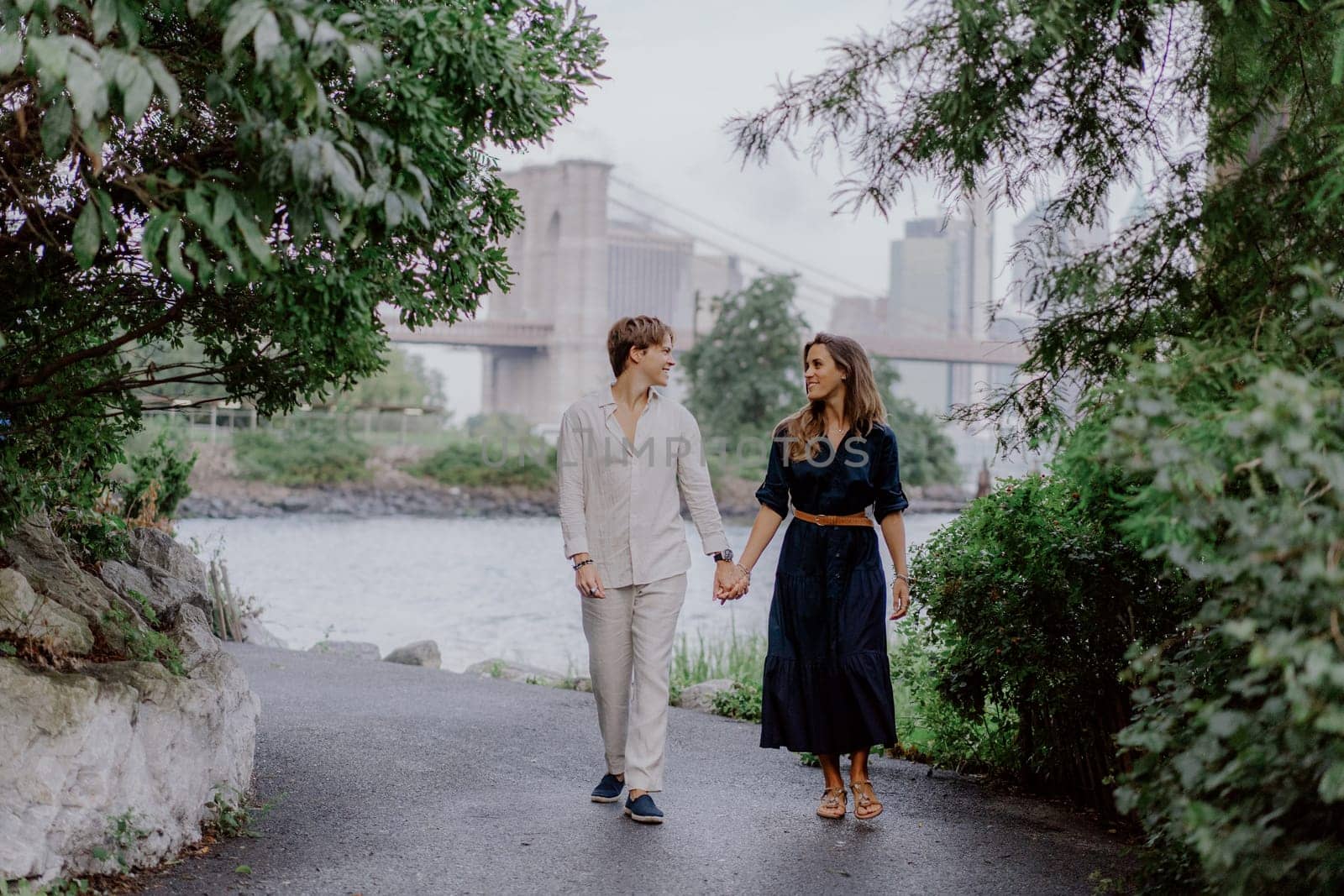 A romantic scene of a couple strolling hand in hand along a waterfront, surrounded by lush greenery and tranquility. Brooklyn bridge in New York.