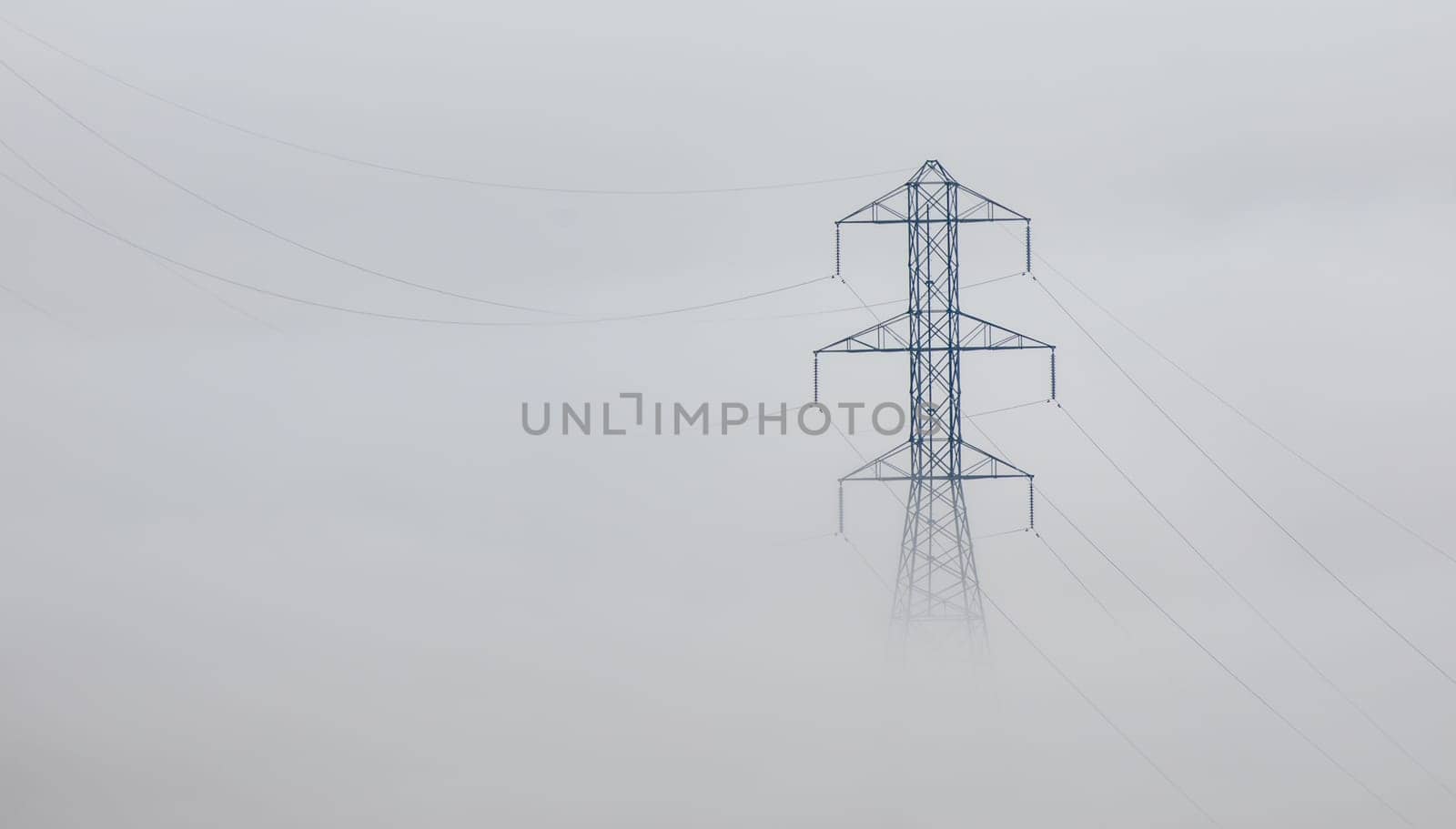 A large metal electricity pylon stands tall amidst a thick fog. Power lines stretch across the misty landscape. No people are visible.