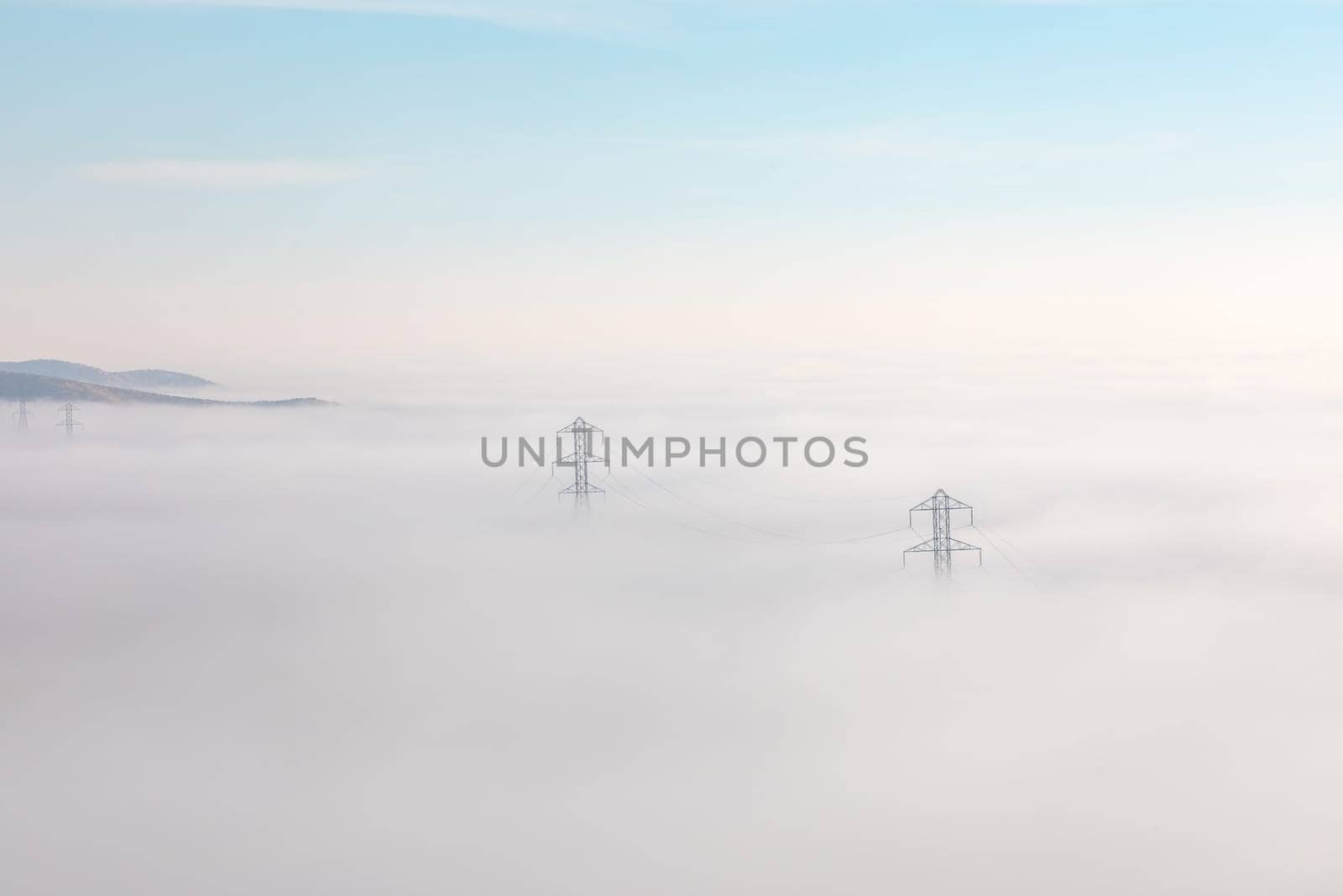 A landscape view of power lines stretching across a thick layer of fog. The fog obscures the terrain, with only the tops of hills visible in the distance