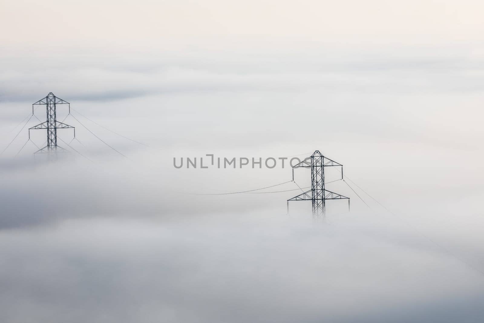 Power lines stretch through a thick fog. Metal pylons pierce the misty landscape, carrying electricity unseen. The fog obscures the ground below, highlighting the infrastructure above.