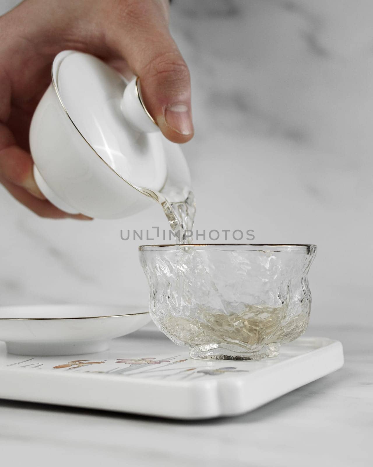 A gentle hand expertly pours steaming tea from a classic white teapot into a beautifully crafted glass cup