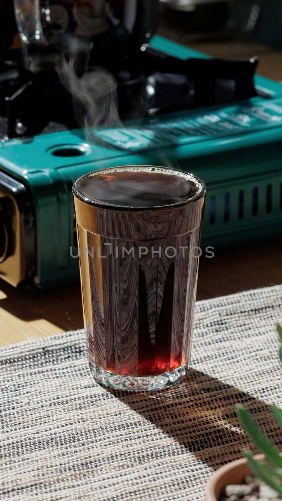 A steaming glass filled with dark beverage rests on a beautifully textured surface close to a gas stove