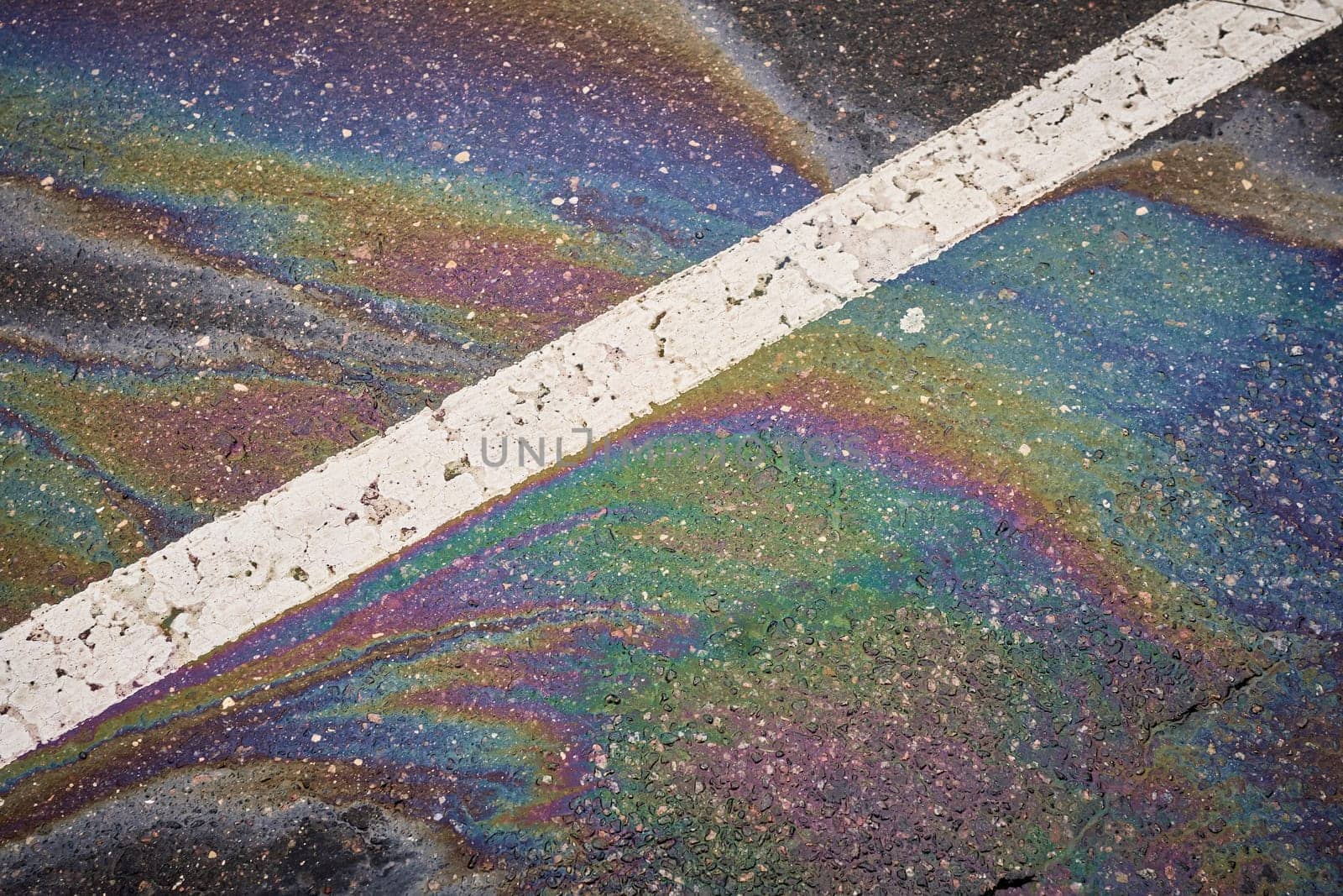 Vibrant oil or gasoline spill producing a rainbow-like sheen on a dark asphalt road, accented by a white traffic line