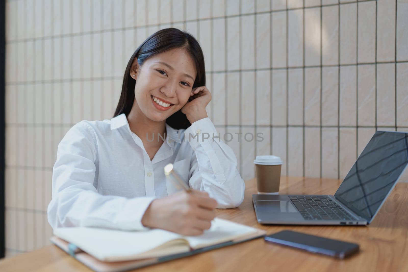 A young woman is happily engaged in her work, using a laptop and jotting down notes, surrounded by a warm and modern office atmosphere filled with wooden elements.