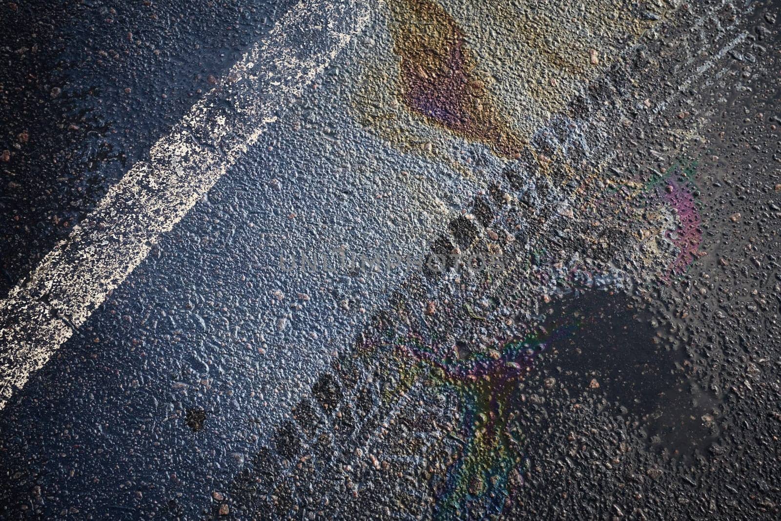 Gasoline rainbow spill reflecting on wet asphalt road surface with white stripe.