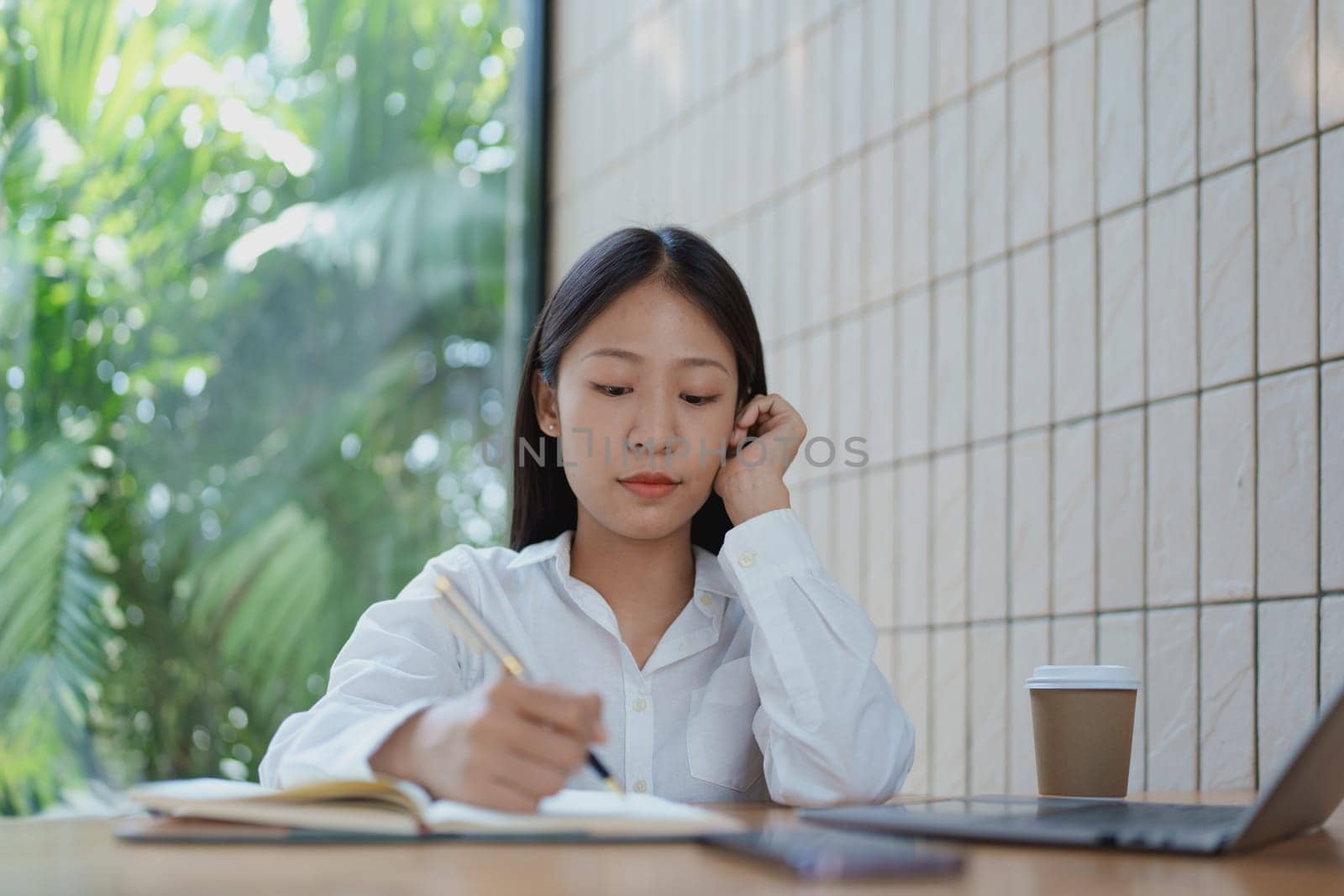 A focused woman writes in her notebook at a cozy wooden table, enjoying a coffee while surrounded by a serene and bright atmosphere filled with greenery.
