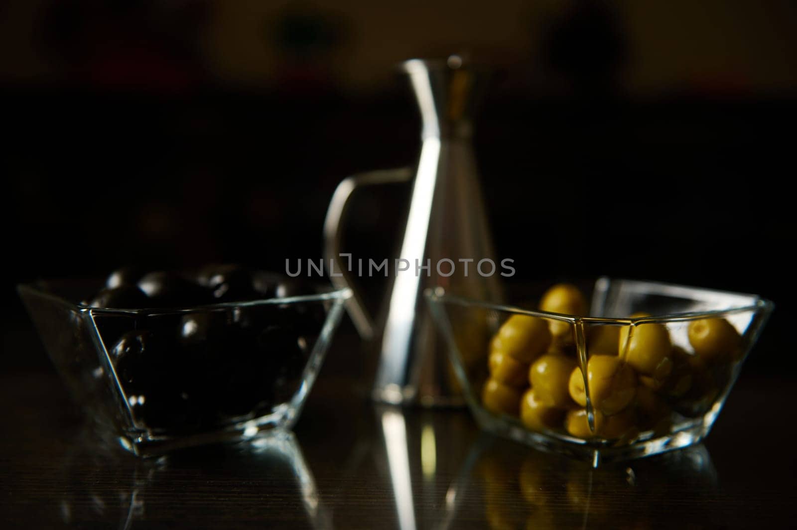 A close-up of two glass bowls containing black and green olives, with a metallic pitcher in the background on a reflective dark table.