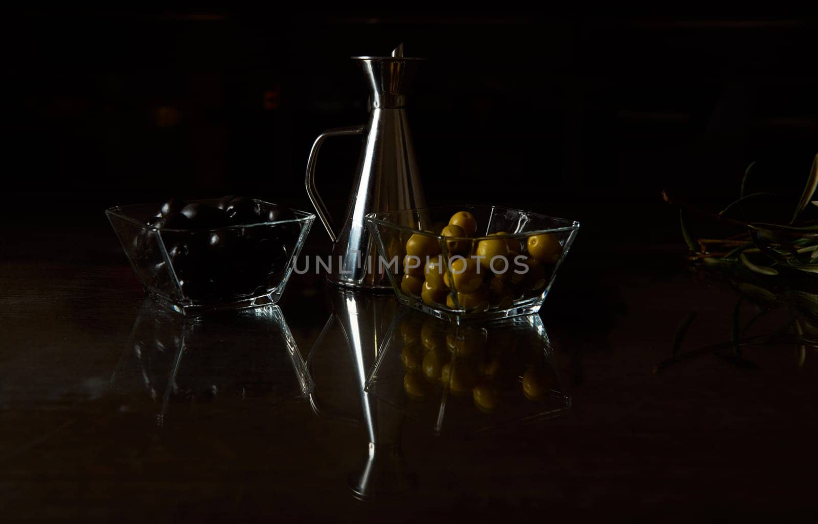 Dark, moody setup featuring black and green olives in glass bowls beside a silver oil can.