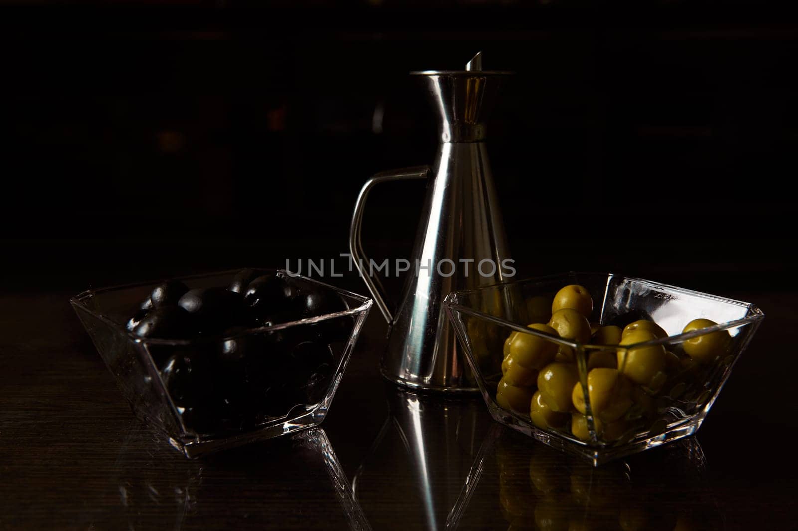 Close-up of dark and moody presentation featuring black and green olives with an olive oil jug. Ideal for culinary themes.