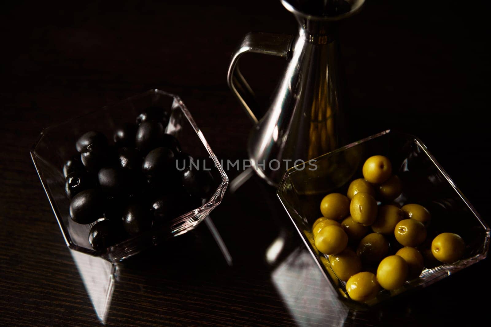 A sophisticated culinary setting featuring black and green olives in glass bowls accompanied by a shiny silver oil jug, highlighting a gourmet kitchen atmosphere.