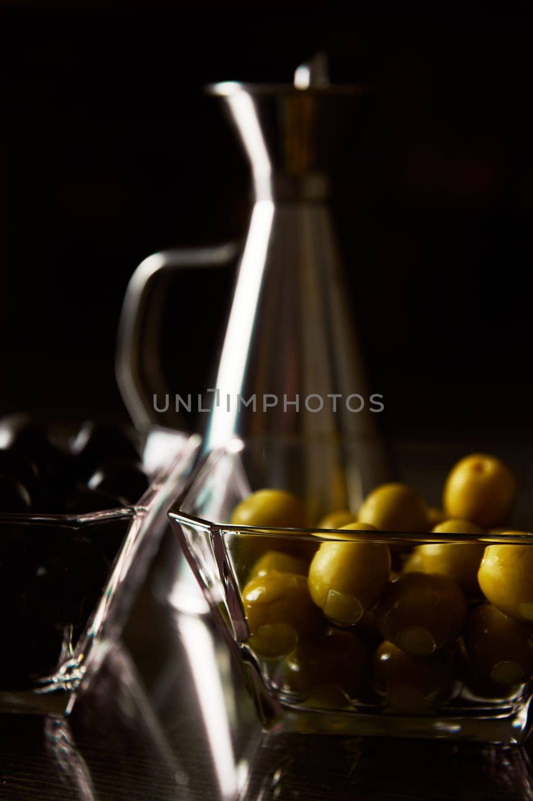Close-up of glass bowls containing green and black olives, paired with a silver oil cruet, under dramatic lighting.