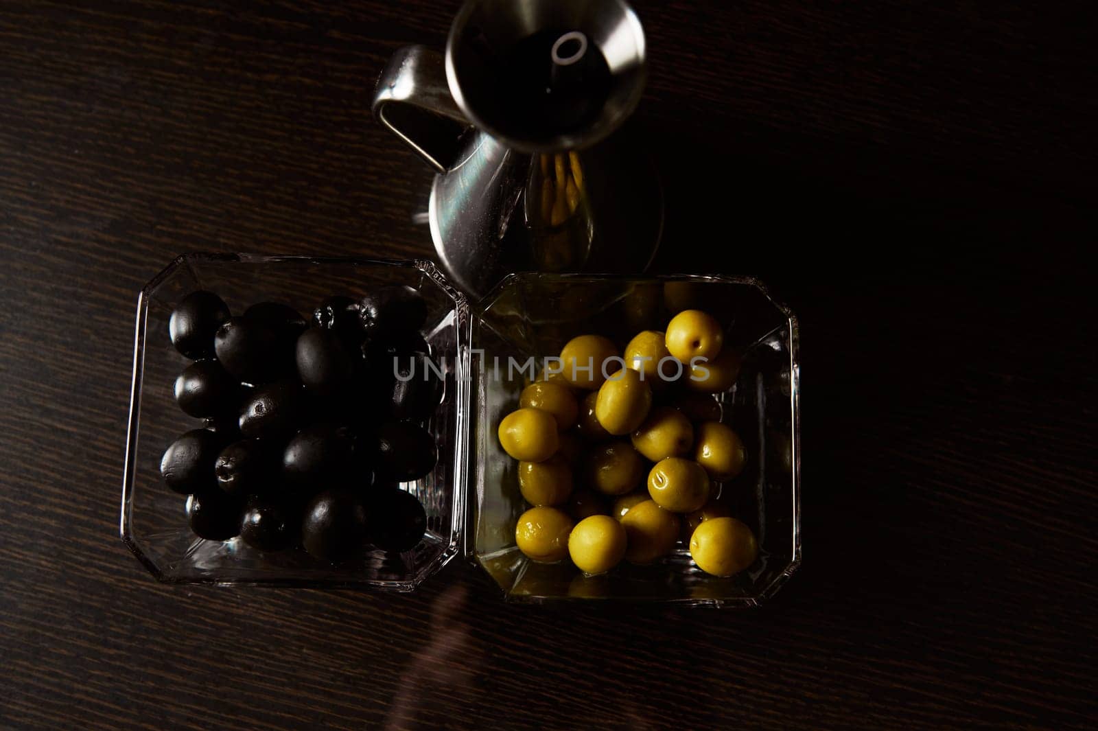 Close-up of black and green olives in clear glass bowls beside a shiny silver oil jug on a dark wooden surface.
