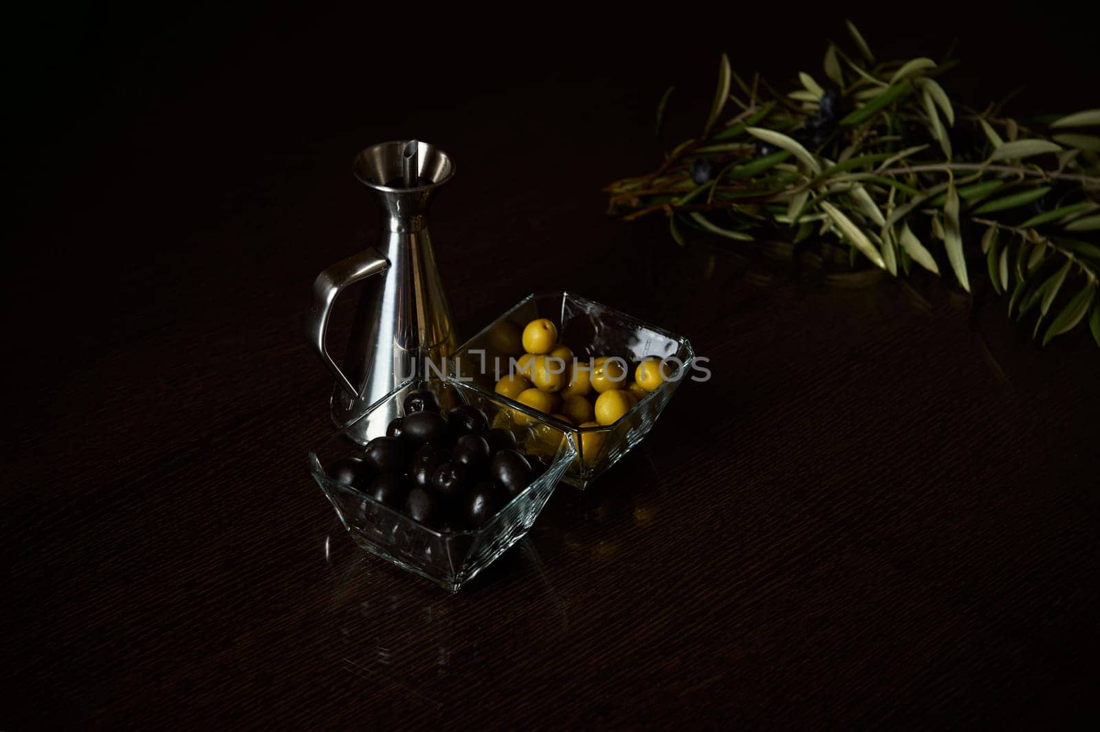 Fresh olive branches next to glass bowls of olives and a metal oil jug on a dark table.