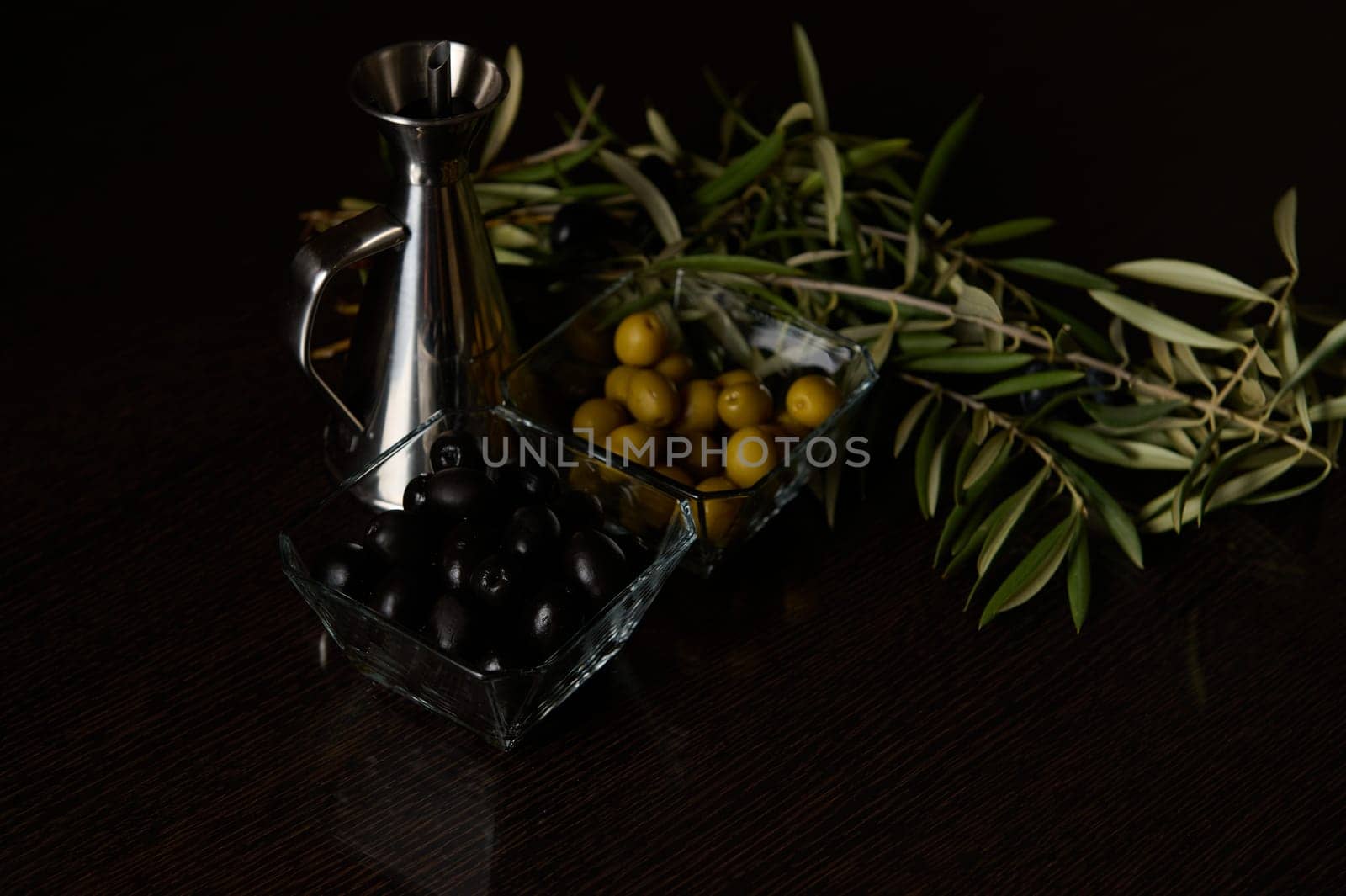 A sophisticated display featuring black and green olives, an olive sprig, and a metallic oil dispenser on a dark surface.