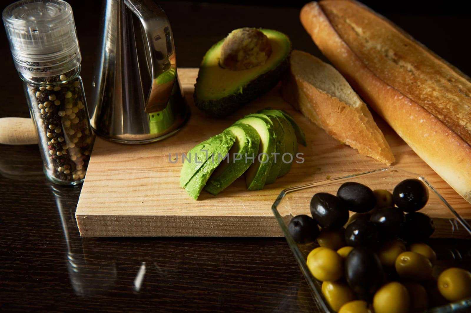 Sliced avocado, black and green olives, and bread on a wooden board create a rustic display. A pepper grinder and oil dispenser complete this culinary scene showcasing natural ingredients.
