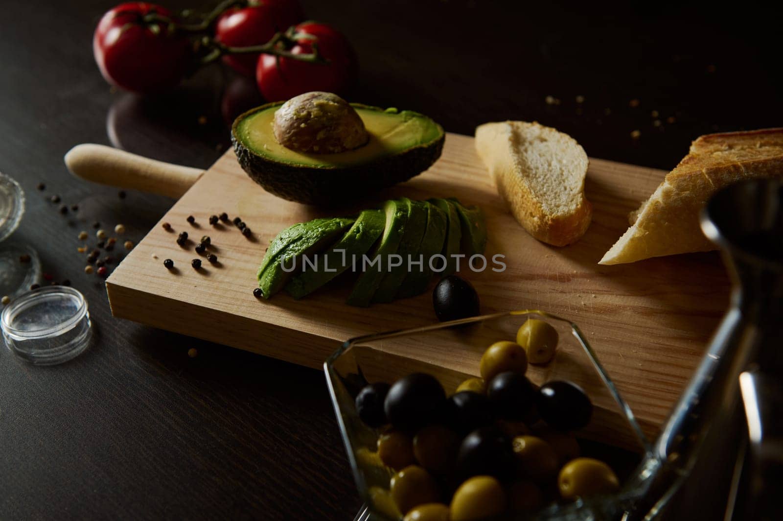 A rustic kitchen scene featuring sliced avocado, bread, olives, and ripe tomatoes on a dark surface.