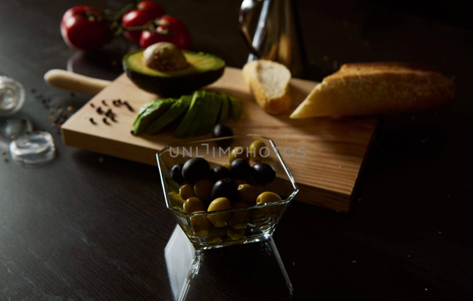 A selection of fresh olives and avocado slices on a cutting board with bread and tomatoes. Prepared for a healthy snack or appetizer on a dark, moody kitchen surface.