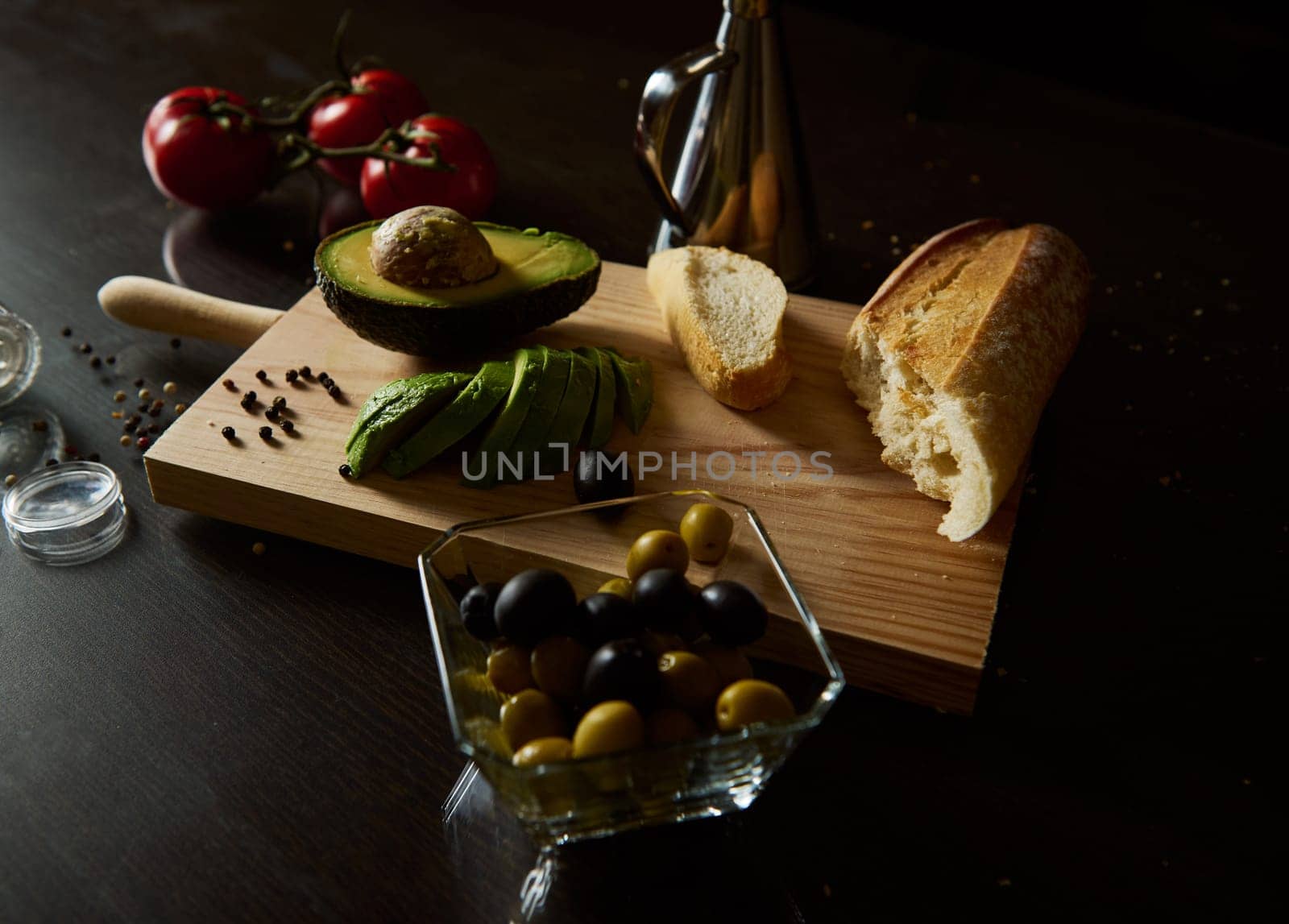 A rustic kitchen setup with sliced avocado, tomatoes, olives, and bread on a wooden board. Ideal for cooking, food styling, and healthy eating concepts in a Mediterranean atmosphere.