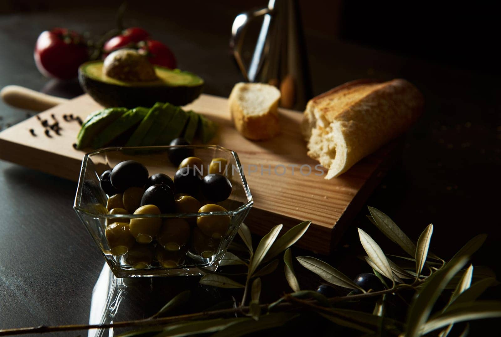 A vibrant display of fresh Mediterranean ingredients including olives, avocado, bread, and tomatoes. The warm, rustic setting highlights a wooden cutting board and a bowl of olives.