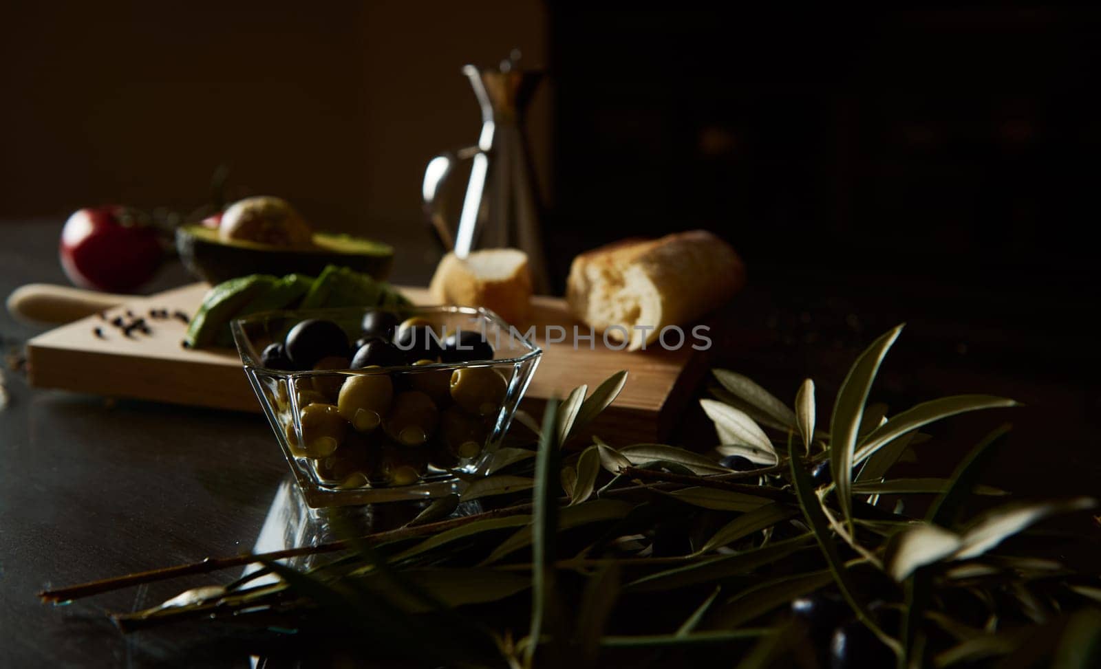 A close-up of olives and a sliced baguette on a rustic kitchen counter, evoking Mediterranean cuisine. Fresh vegetables and olive branches add to the authentic and inviting cooking scene.