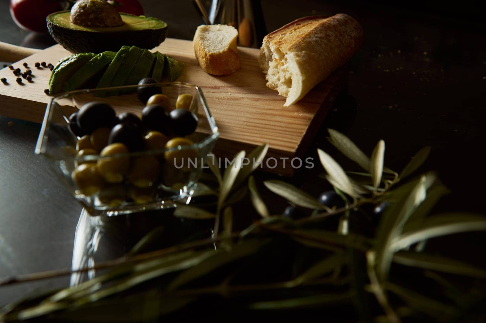 A rustic composition featuring fresh olives, sliced avocado, and baguette, evoking Mediterranean flavors.