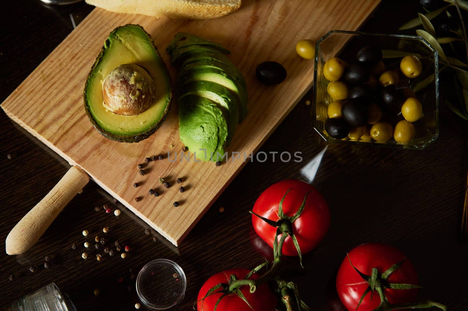 Sliced avocado and whole tomatoes on a board surrounded by olives and spices.
