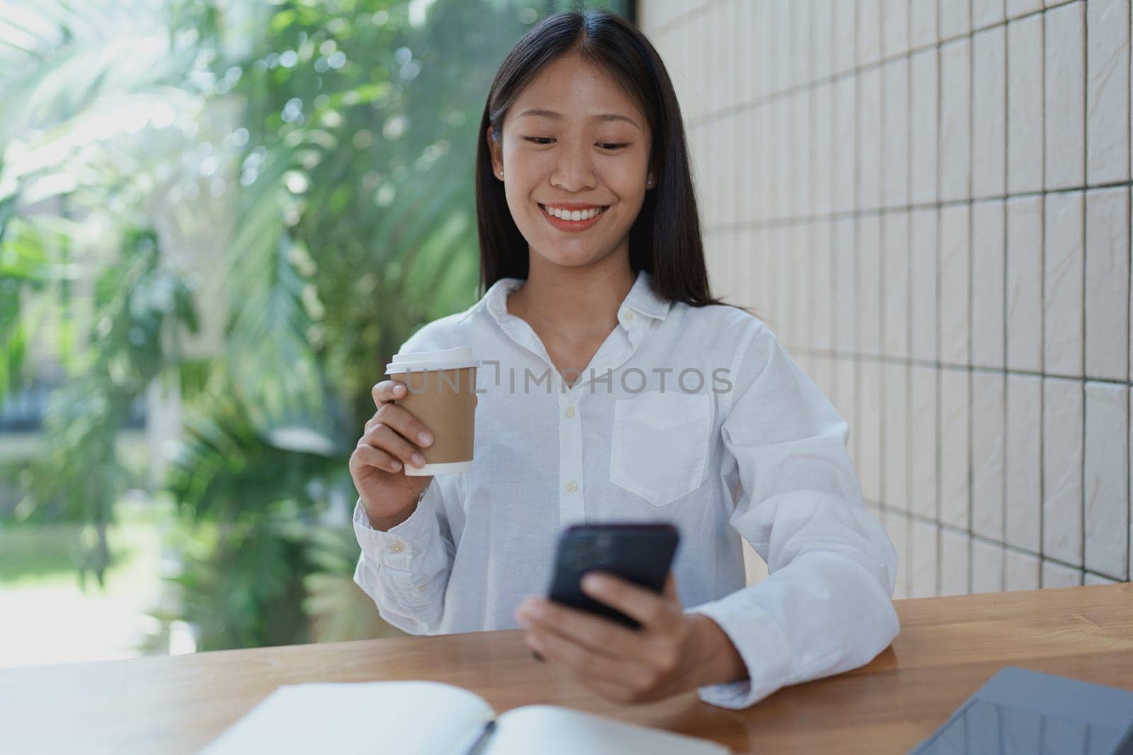 A cheerful young woman sits at a wooden table, sipping coffee and using her smartphone. The setting is bright and filled with greenery, reflecting a modern, relaxed lifestyle.