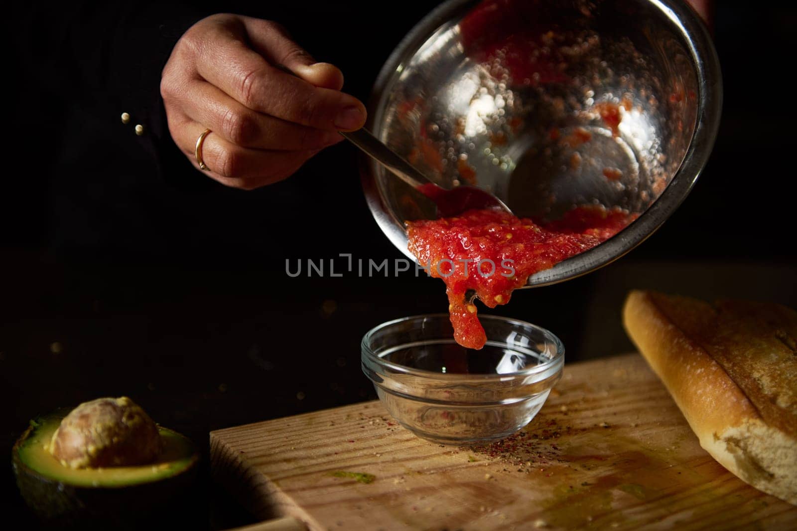 A person pours freshly made tomato sauce into a glass bowl. Avocado and bread slices are on the cutting board, ready to serve.