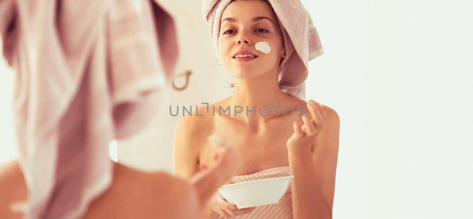 A young girl in a towel on her head and body stands near the mirror in the bathroom and applies a clay mask to her face, the woman takes care of her health and beauty.