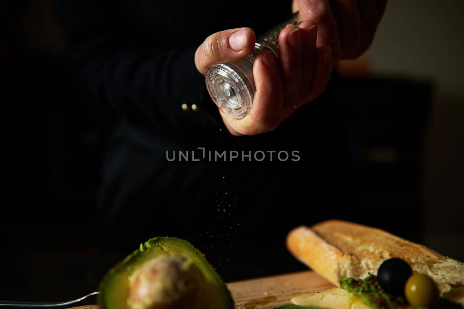 A person seasoning fresh avocado with salt, highlighting kitchen preparation. The scene includes bread and olives, emphasizing culinary art and home cooking atmosphere.